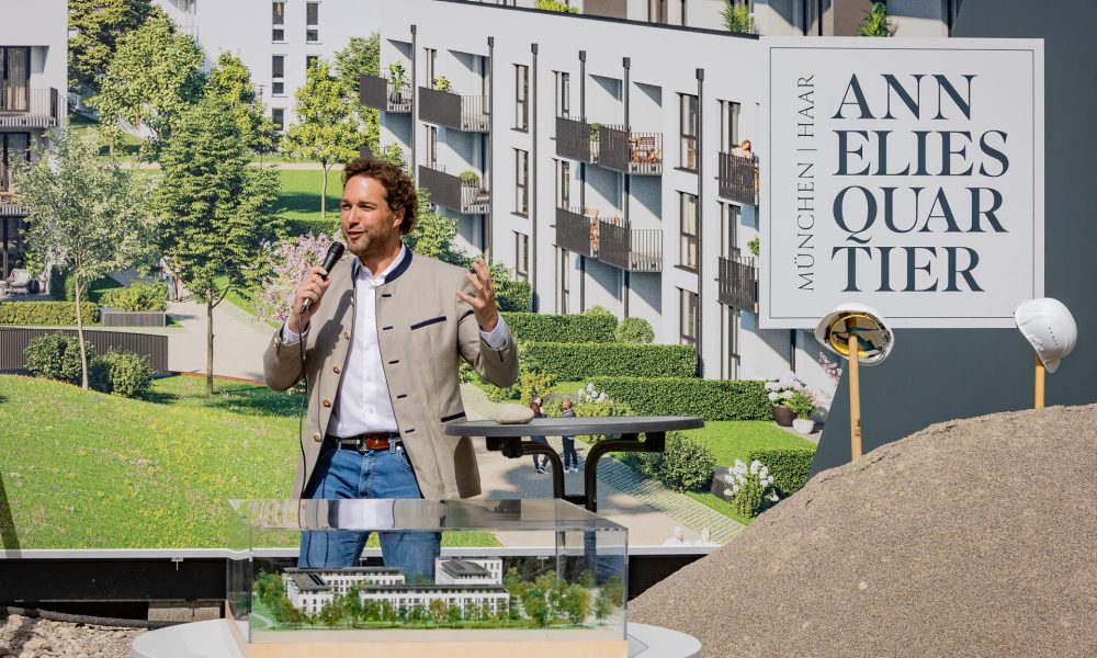 Bürgermeister Dr. Andreas Bukowski beim Spatenstich präsentiert Annelies Quartier mit Mikrofon und Werbeplane im Hintergrund